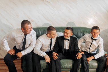 Dört adam kanepede oturuyor, hepsi siyah takım elbiseli ve kravatlı. Gülümsüyorlar ve birbirlerinin arkadaşlığından hoşlanıyor gibi görünüyorlar. Sahne neşeli ve dostane.