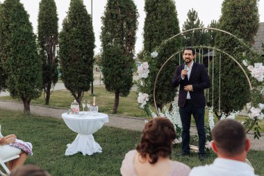 Bir adam düğün kemerinin önünde mikrofona konuşuyor. Bir grup insan onu izliyor, bazıları sandalyede oturuyor. Sahne, arka planda ağaçlar olan bir parkta geçiyor.