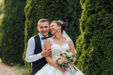 Bir gelin ve damat çitin önünde fotoğraf için poz veriyorlar. Gelin bir buket çiçek tutuyor ve damat kravat takıyor. İkisi de gülümsüyor ve mutlu görünüyorlar.