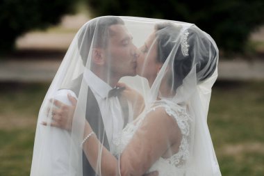 Bir gelin ve damat peçenin altında öpüşüyor. Gelin beyaz bir elbise giyiyor ve damat siyah bir takım giyiyor.