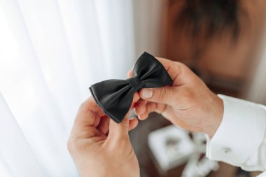 Bir adam elinde siyah bir papyon tutuyor. Papyon parlak ve iyi yapılmış görünüyor. Adam resmi bir etkinliğe hazırlanıyor gibi görünüyor. Düğün ya da iş toplantısı gibi.