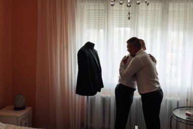 İki adam siyah takım elbiseli bir mankenin önünde birbirlerine sarılıyorlar. Sahne samimi ve sıcak, erkeklerin birbirlerine sarıldığı bir an.