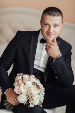 Siyah takım elbiseli ve beyaz kravatlı bir adam elinde bir buket çiçek tutuyor.