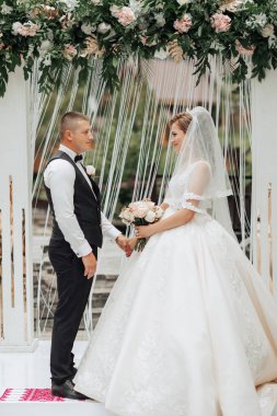 Bir gelin ve damat beyaz bir kemerin altında duruyor. Gelin elinde bir buket çiçek tutuyor.