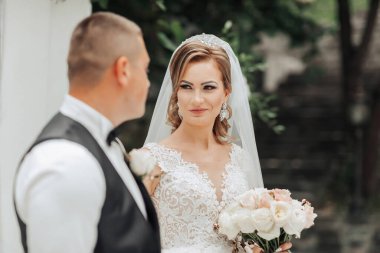 Bir gelin ve damat beyaz bir binanın önünde duruyor. Gelin elinde bir buket çiçek tutuyor.