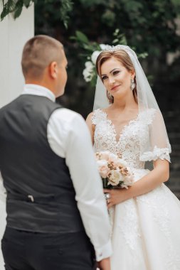 Bir gelin ve damat beyaz bir kemerin önünde duruyor. Gelin bir buket çiçek tutuyor ve damat bir buket çiçek tutuyor.