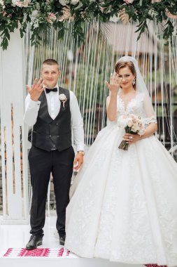 Gelin ve damat beyaz bir kemerin önünde duruyorlar, gelin elinde buketle