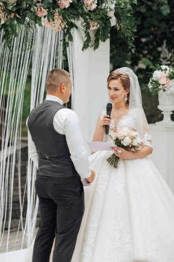 Bir gelin ve damat beyaz bir kemerin önünde duruyor. Gelin bir mikrofon tutuyor ve damat da bir dosya tutuyor.