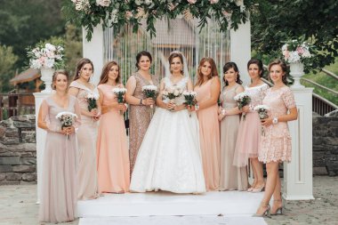 Bir grup kadın düğün fotoğrafı için poz veriyor. Gelin beyaz bir elbise giyiyor ve nedimeleriyle çevrili. Kadınların hepsi pembe giyinmiş ve ellerinde çiçekler var.