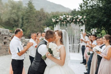 Bir gelin ve damat kalabalığın önünde öpüşüyor. Gelin beyaz bir elbise giyiyor ve damat siyah bir yelek giyiyor. Düğün konukları yeni evliler için alkış ve tezahürat yapıyor.