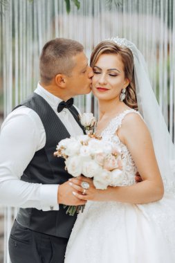 Bir gelin ve damat bir buket çiçek tutarken birbirlerini öpüyorlar. Gelin beyaz bir elbise giyiyor ve damat siyah bir takım elbise giyiyor. Sahne bir düğün.