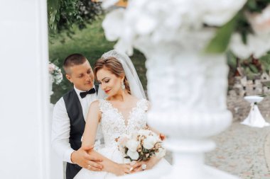 Bir gelin ve damat beyaz bir vazonun önünde fotoğraf için poz veriyorlar. Gelin elinde bir buket çiçek tutuyor.