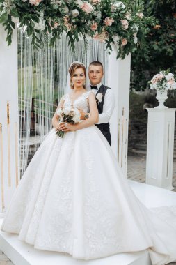 Bir gelin ve damat beyaz bir kemerin önünde poz veriyorlar. Gelin beyaz bir elbise giyiyor ve damat siyah bir yelek giyerken elinde bir buket tutuyor. Sahne bir bahçede geçiyor.