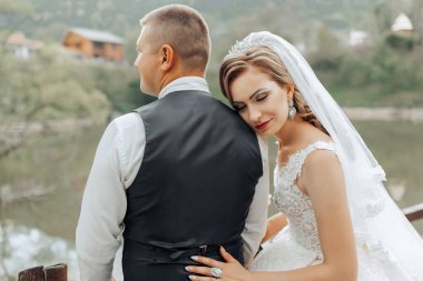 Bir gelin ve damat, köprünün üzerinde bir su birikintisine bakan bir resim için poz veriyorlar. Gelin beyaz bir elbise ve duvak giyerken damat takım elbise ve kravat takıyor. İkisi de gülümsüyor.