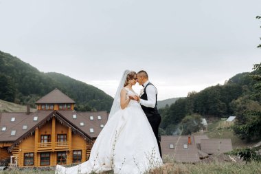 Gelin ve damat, dağları gören bir evin önünde kucaklaşır.
