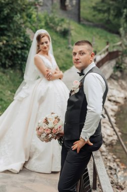 Bir gelin ve damat, köprüde fotoğraf çektirmek için poz veriyor. Gelin beyaz bir elbise giyiyor ve damat siyah bir yelek giyiyor. Sahne mutlu ve kutlama dolu, çünkü çift evleniyor.