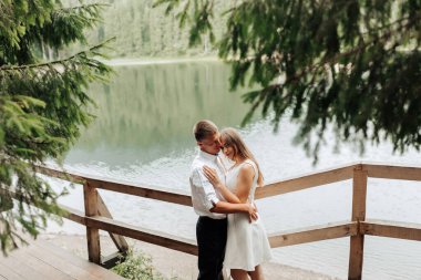 Göle bakan ahşap bir köprüde beyaz elbiseli bir kadınla duran bir çift var.