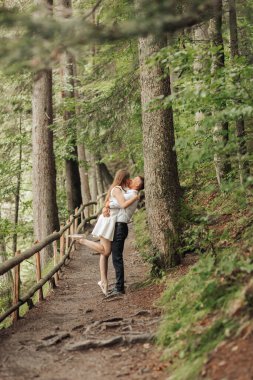 Bir çift, kadının arkasında duran adamla ormanda kucaklaşıyorlar. Sahne romantik ve huzurlu.