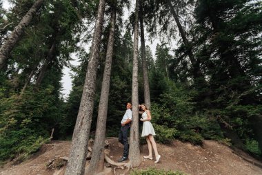 Bir çift ormanda bir ağacın önünde poz veriyor. Adam beyaz bir gömlek giyiyor ve kadın beyaz bir elbise giyiyor. Sahne romantik ve huzurlu.