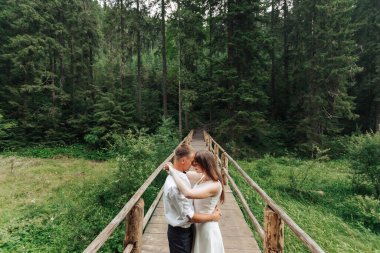 Bir çift ormanda tahta bir köprüde kucaklaşıyor. Sahne romantik ve huzurlu.