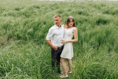 Bir adam ve bir kadın uzun otların arasında duruyorlar. Adam beyaz bir gömlek giyiyor ve kadın beyaz bir elbise giyiyor. İkisi de gülümsüyor ve kameraya bakıyor.