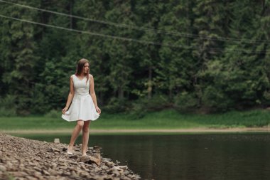 Beyaz elbiseli bir kadın gölün yanındaki kayalık bir sahilde duruyor. Sahne sakin ve huzurlu, kadın denize bakıyor.