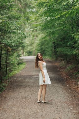 Bir kadın ormandaki toprak bir yolda duruyor. Beyaz bir elbise giyiyor ve gülümsüyor.