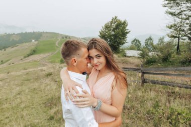 Arka planda çitleri olan bir çift tarlada kucaklaşıyor. Kadın pembe bir elbise giyiyor ve adam beyaz bir gömlek giyiyor. Sahne romantik ve samimi.