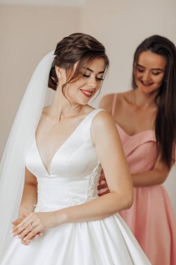 Bir gelin nedimesiyle düğününe hazırlanıyor. Gelin beyaz bir elbise ve duvak takıyor. Sahne mutlu ve kutlama dolu.