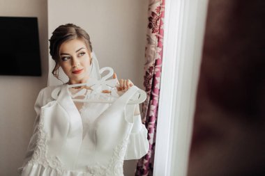 Bir kadın pencerenin önünde beyaz bir elbise tutuyor. Elbise askıda asılı duruyor ve kadın ona bakıyor. Sahne beklenti ve heyecandan ibarettir.