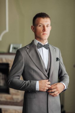 Takım elbiseli ve kravatlı bir adam şöminenin önünde duruyor. Gri bir takım elbise ve siyah bir papyon giyiyor.