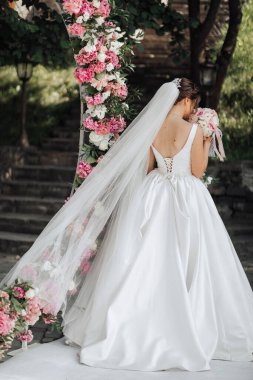 Beyaz elbiseli bir kadın pembe kemer yolunun önünde duruyor. Elinde bir buket çiçek ve duvak tutuyor. Sahne muhtemelen bir düğün ya da özel bir olay.