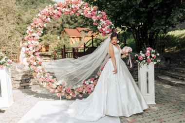 Pembe çiçeklerle büyük bir kemer yolunun önünde bir gelin duruyor. Beyaz bir elbise giyiyor ve elinde bir buket tutuyor. Sahne arka planda bir ev olan bir bahçede geçiyor.