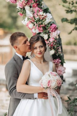 Bir gelin ve damat pembe bir kemerin önünde poz veriyorlar. Gelin bir buket çiçek tutuyor ve damat onu yanağından öpüyor. Sahne romantik ve mutlu.