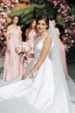 Bir gelin beyaz bir elbise giyiyor ve elinde bir buket pembe çiçek tutuyor. Pembe bir kemerin önündeki beyaz halıda duruyor.