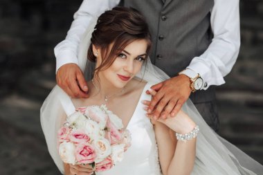 Bir gelin ve damat bir buket çiçekli fotoğraf için poz veriyorlar. Gelin beyaz bir elbise giyiyor ve damat gri bir yelek giyiyor. Sahne mutlu ve romantik.