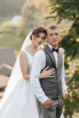 Güneşli bir günde bir gelin ve damat fotoğraf için poz veriyor. Gelin beyaz bir elbise giyiyor ve damat siyah bir takım elbise giyiyor. İkisi de gülümsüyor ve kameraya bakıyor.