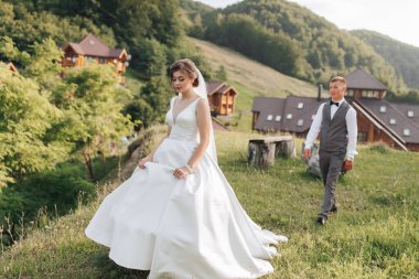 Bir gelin ve damat arka planda dağların güzel manzarasıyla çimenli bir yamaçta yürüyorlar. Gelin beyaz bir elbise ve duvak takarken damat takım elbise ve kravat takıyor.