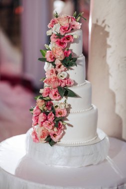 Üzerinde pembe çiçekler olan beyaz bir pasta. Pasta uzun ve dört katlı. Çiçekler, pastanın kenarlarından dökülüyor gibi görünecek şekilde düzenlenmiş.