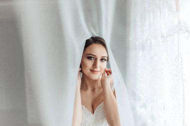 Bir kadın peçe takıyor ve perdenin önünde duruyor. Yüzünde bir gülümsemeyle kameraya bakıyor.