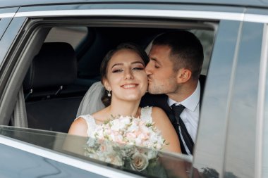 Bir gelin ve damat arabada öpüşüyor. Gelin elinde bir buket çiçek tutuyor.