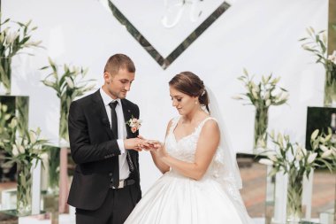 Bir gelin ve damat büyük bir aynanın önünde evleniyorlar. Gelin beyaz bir elbise giyiyor ve damat siyah bir takım elbise giyiyor. Onlar büyük bir çiçek vazosunun önünde duruyorlar.