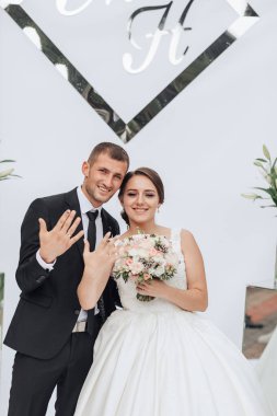 Bir gelin ve damat üzerinde F ve H harfleri olan beyaz bir duvarın önünde poz veriyorlar.