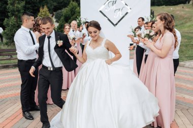Bir gelin ve damat bir grup insanın önünde dans ediyor. Gelin beyaz bir elbise giyiyor ve damat siyah bir takım elbise giyiyor. Misafirler yeni evliler için alkış ve tezahürat yapıyorlar.