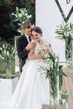 Bir gelin ve damat beyaz bir kemer yolunun önünde poz veriyorlar. Gelin bir buket çiçek tutuyor ve damat kravat takıyor. Sahne muhtemelen bir düğün ya da resmi bir etkinlik.