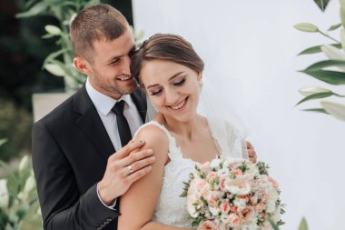Bir gelin ve damat beyaz bir arkaplan önünde sarılıp gülümsüyor. Gelin elinde bir buket çiçek tutuyor.