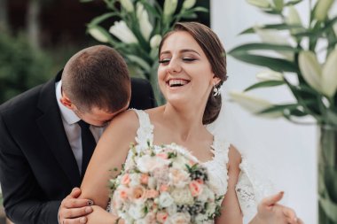 Bir gelin ve damat birbirlerine sarılıp öpüşüyorlar. Gelin elinde bir buket çiçek tutuyor.