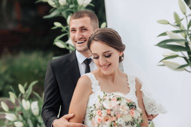 Bir gelin ve damat bir buket çiçekli fotoğraf için poz veriyorlar. Gelin beyaz bir elbise giyiyor ve damat bir takım elbise giyiyor. Sahne mutlu ve kutlama dolu.