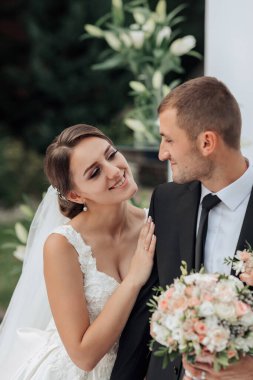 Bir gelin ve damat bir buket çiçekli fotoğraf için poz veriyorlar. Gelin beyaz bir elbise giyiyor ve damat siyah bir takım elbise giyiyor. Sahne mutlu ve romantik.