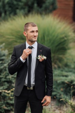 Takım elbiseli ve kravatlı bir adam yakasında çiçekle çalıların önünde duruyor.
